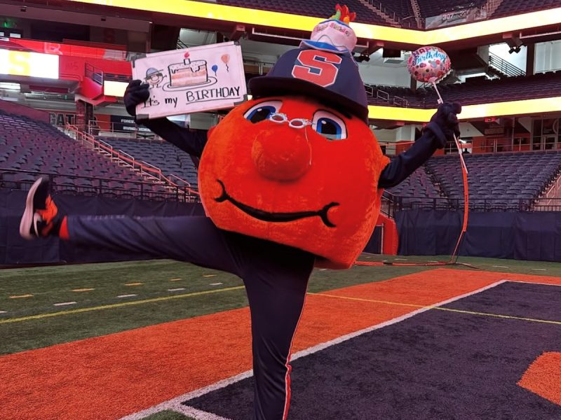 Otto the Orange celebrates on the JMA Wireless Dome field wearing a birthday party hat, holding an "It's my BIRTHDAY" sign in one hand and a Happy Birthday balloon in the other, with one leg kicked up.