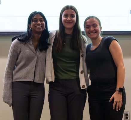Three students standing together and smiling in front of a projected presentation screen