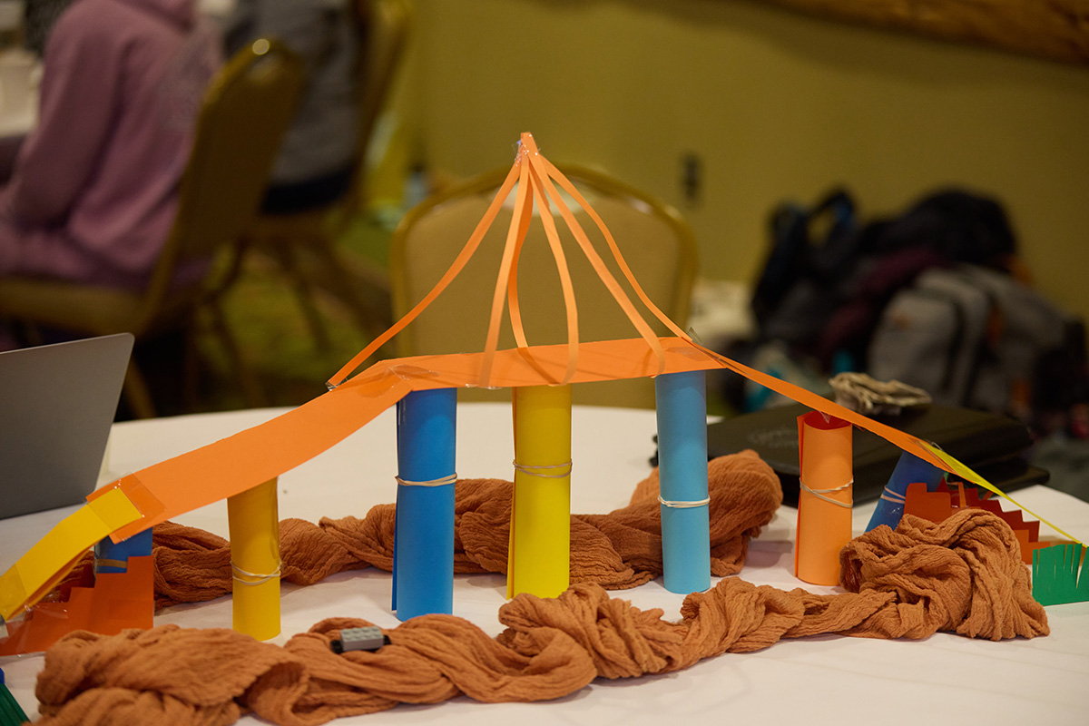 A handmade bridge model constructed from colorful rolled paper tubes, orange paper strips, tape, and brown fabric draped around the base, displayed on a table during a group activity.