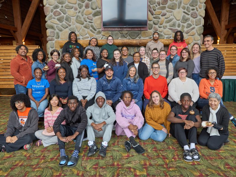 A group of approximately 40 individuals pose together for a photo in a lodge-style room with a stone fireplace, some wearing Syracuse gear and several holding small golden pumpkins.