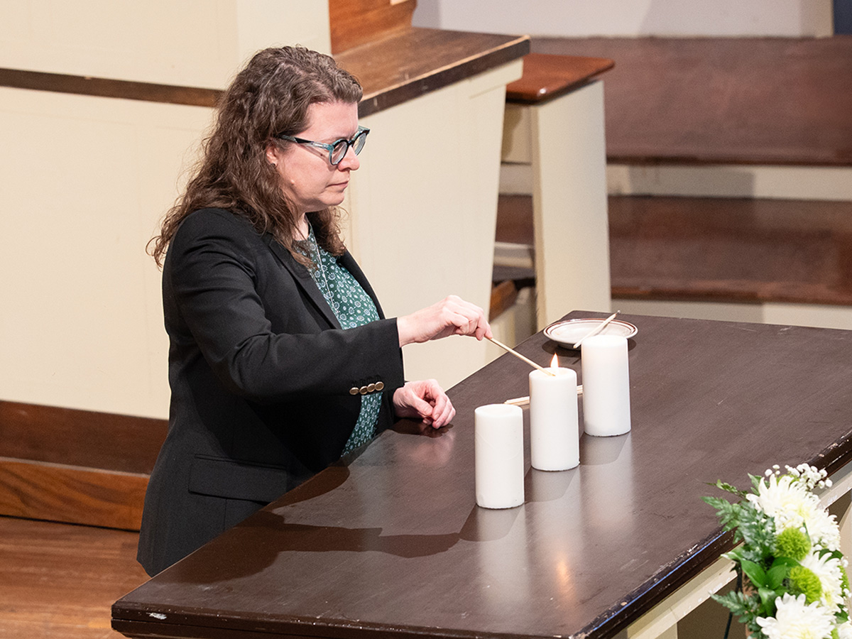 A woman with curly brown hair and glasses lights white pillar candles on a wooden table in a church, using a long match or candle lighter, with a floral arrangement visible in the foreground.