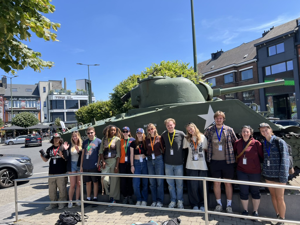 A group of about a dozen students poses in front of a green Sherman tank on a sunny city street.