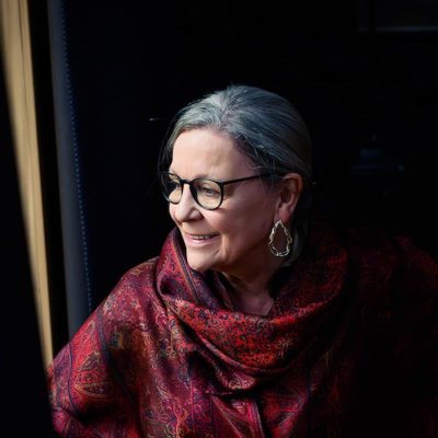 Person seated near a window wearing a patterned red shawl, softly lit by natural light against a dark interior background
