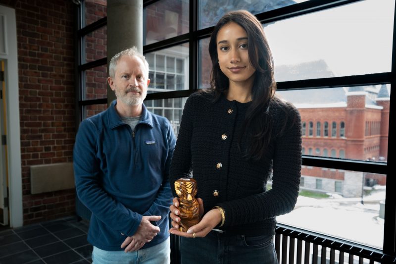 A young person with long dark hair holds a small carved figurine while standing adjacent to a middle-aged man with gray hair and a beard.