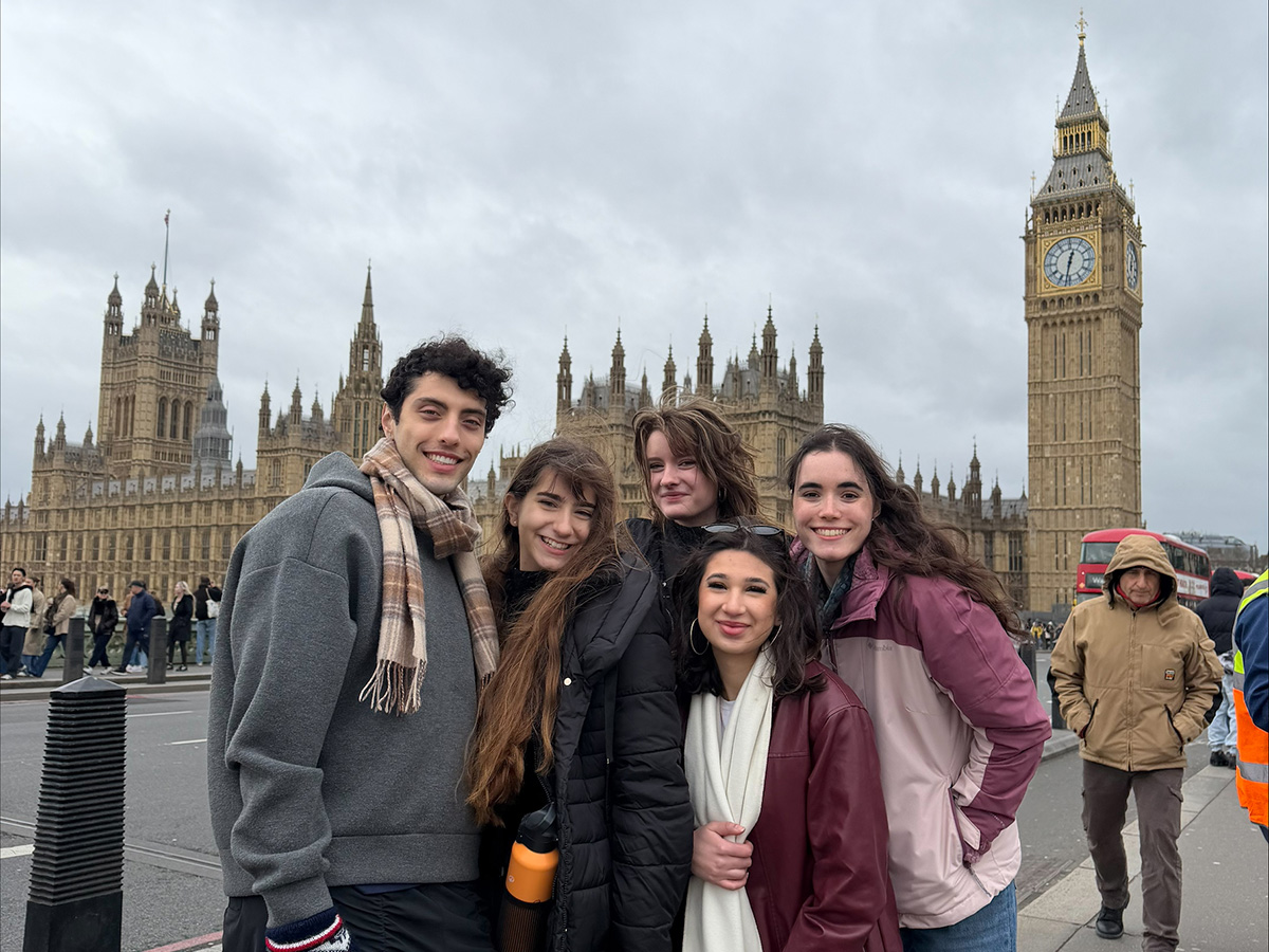 Five smiling students pose together on Westminster Bridge in London, with the Houses of Parliament and Big Ben visible in the background on an overcast day.