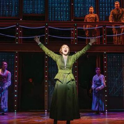 A performer in a green dress sings center stage with arms raised, surrounded by cast members on a two-level set.