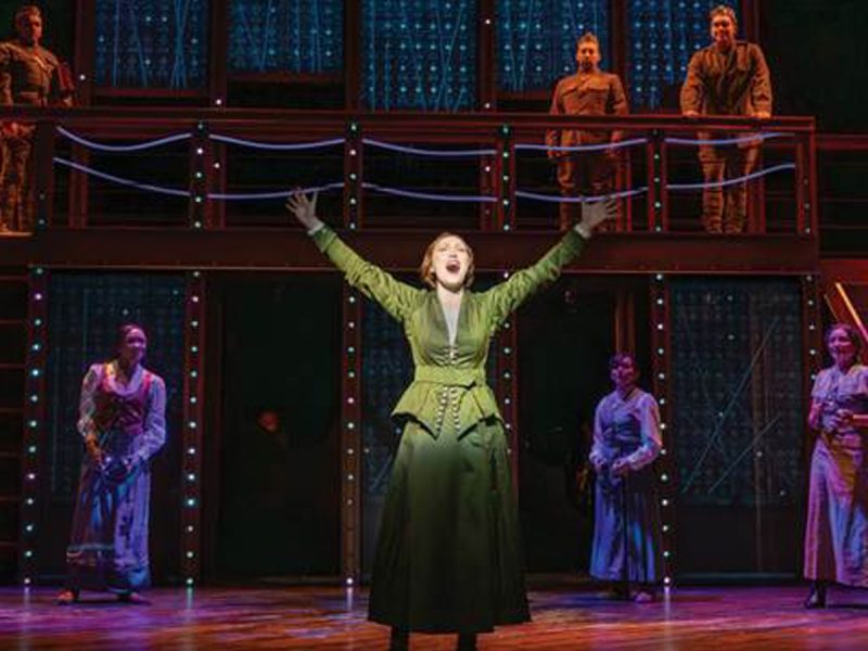 A performer in a green dress sings center stage with arms raised, surrounded by cast members on a two-level set.