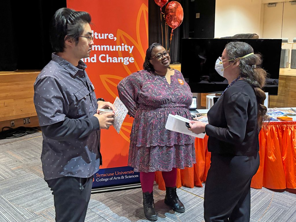 Three people standing and talking; an orange banner that says Culture. Community and Change is in the background.