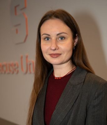 A person with long auburn hair and blue eyes looks composed and is wearing a grey striped blazer and maroon top.