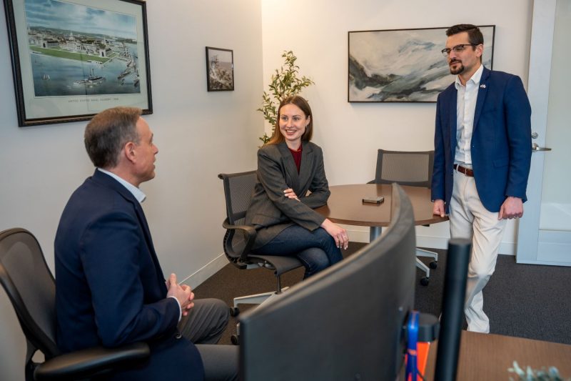 Three people engage in conversation in a professional office setting, with framed artwork and a print of the United States Naval Academy visible on the walls.