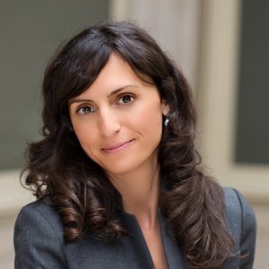 Professional head-and-shoulders portrait of a person with dark, shoulder-length hair wearing a gray blazer against a neutral background.