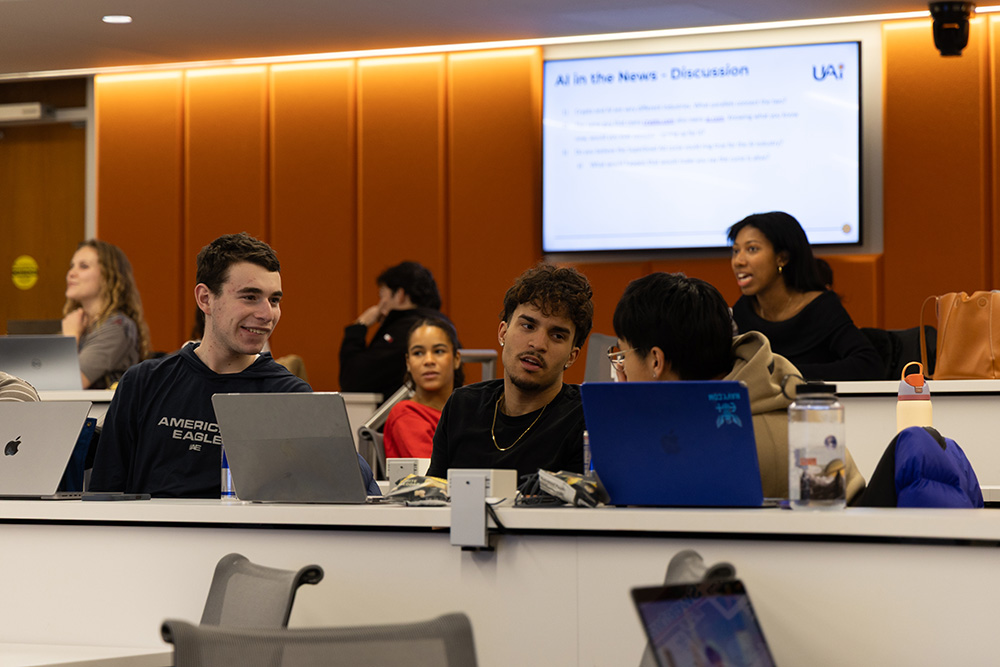 Students seated at classroom desks using laptops during a group discussion, with “AI in the News” displayed on a screen