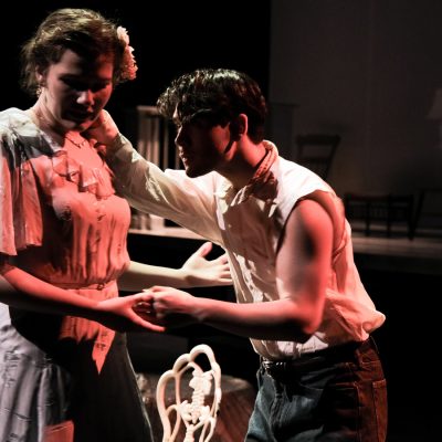 Two student actors perform an intimate scene on stage, bathed in warm red light.