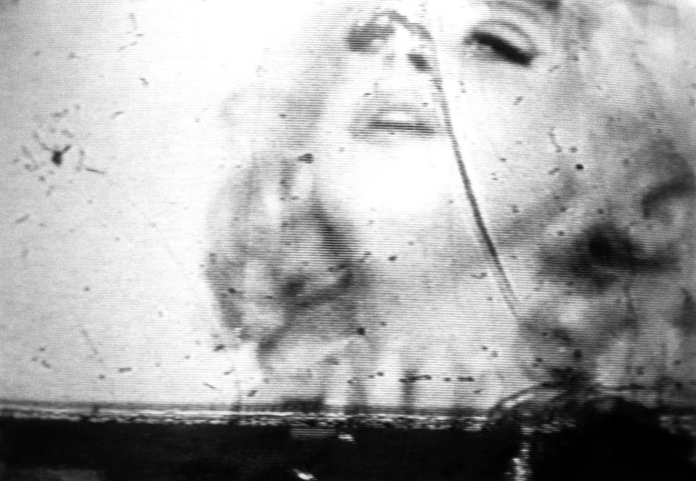 A grainy, distorted black-and-white photograph of a figure's face, in John Waters' "Dirty Divine" (2000), a gelatin silver print gifted to the Syracuse University Art Museum by Nancy Delman Portnoy.