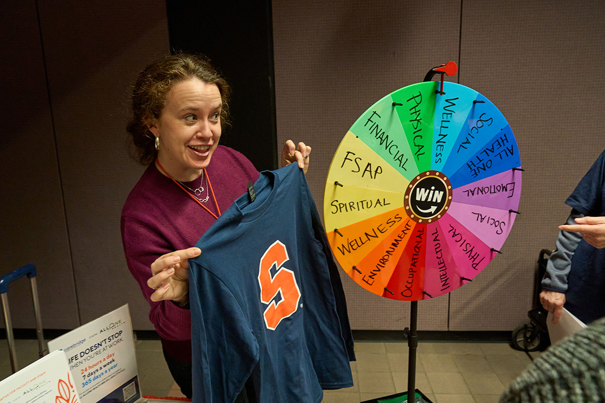 Person holding a Syracuse Block S shirt beside a colorful wellness wheel display