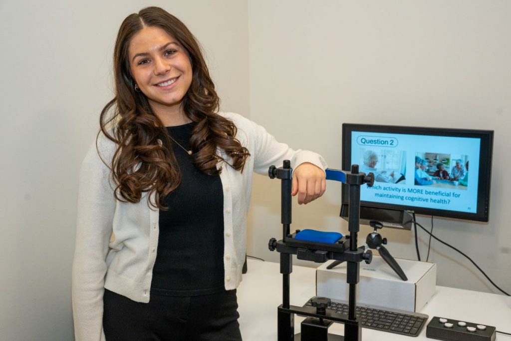 A person with long dark hair smiles while standing next to research equipment in a lab setting. Behind her, a monitor displays a slide reading, "Question 2: Which activity is MORE beneficial for maintaining cognitive health?"