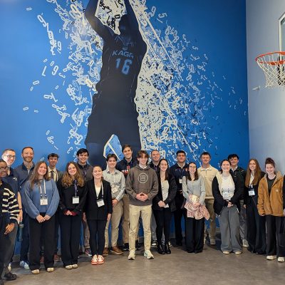 A group of approximately 20 students and professionals pose in front of a large blue mural depicting a basketball player wearing jersey number 16, with a basketball hoop visible on the right wall
