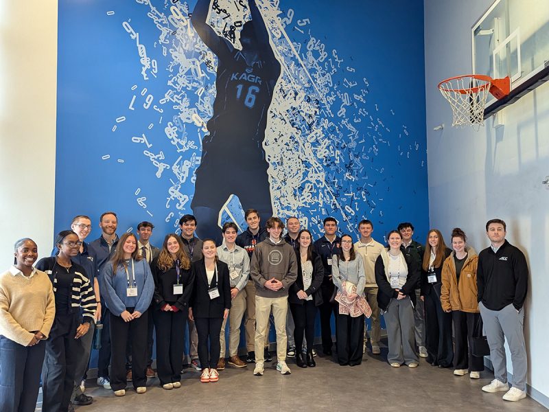 A group of approximately 20 students and professionals pose in front of a large blue mural depicting a basketball player wearing jersey number 16, with a basketball hoop visible on the right wall