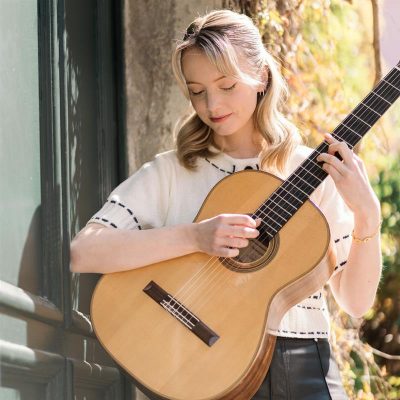 Person playing a classical acoustic guitar outdoors beside a building, wearing a light short‑sleeve top, with sunlight and greenery in the background.
