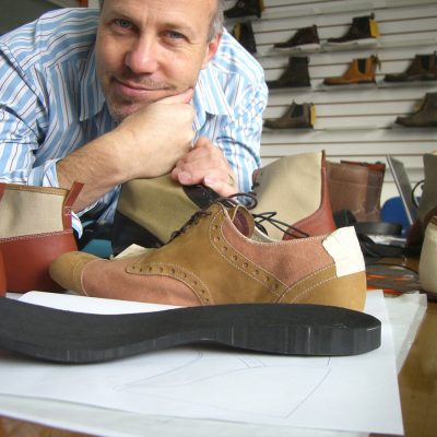 A man leans on a workbench, smiling at the camera with his chin resting on his folded hands. In front of him is a tan canvas and suede brogue-style oxford shoe prototype with a separate black rubber sole on design sketches. Several other boot and shoe prototypes in canvas and leather are visible on the desk and displayed on a wall in the background, suggesting a shoe design studio or workshop.