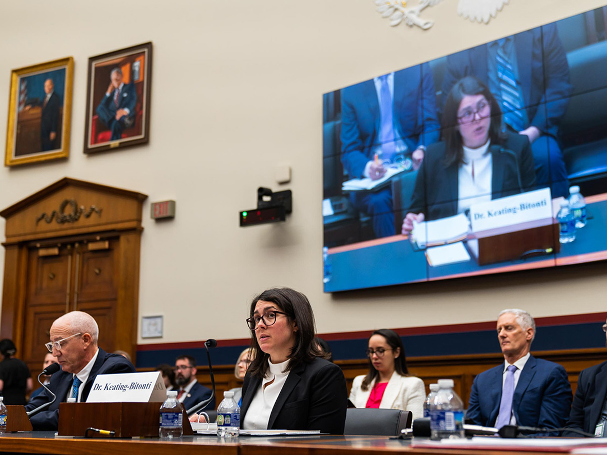 A congressional hearing room where several witnesses are seated at a long wooden testimony table with microphones and water bottles. In the center, a woman identified by a nameplate as Dr. Keating-Bitonti — wearing a black blazer over a white turtleneck and glasses — speaks into a microphone. She is simultaneously displayed on a large video screen mounted on the wall behind the panel. To her left sits an older man in a dark suit with glasses, and to her right sits another man in a dark suit with a blue tie. Official portraits hang on the cream-colored walls in the background.