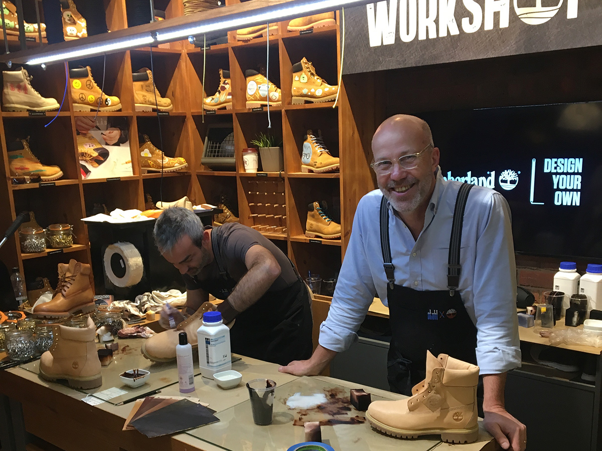 Two men work at a Timberland "Design Your Own" workshop booth. In the foreground, a man with glasses smiles at the camera, wearing a light blue shirt with dark suspenders over a work apron. Beside him, a second man leans over the workbench, focused on crafting a boot. The workbench is covered with tools, dyes, leather pieces, and unfinished tan Timberland boots. Behind them, wooden cubby shelves display numerous customized Timberland boots in various painted designs. A screen in the background reads "Timberland – Design Your Own."