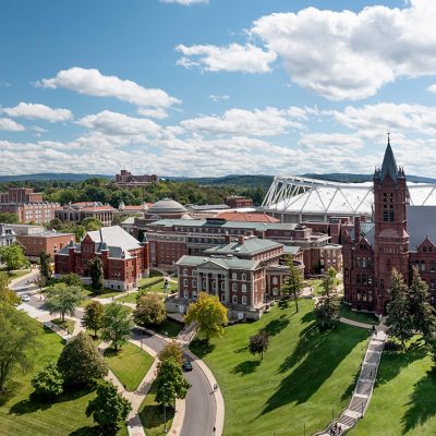 Aerial view of Syracuse University campus showing red-brick buildings, green lawns, Crouse College's tower, and the JMA Wireless Dome under a partly cloudy sky.