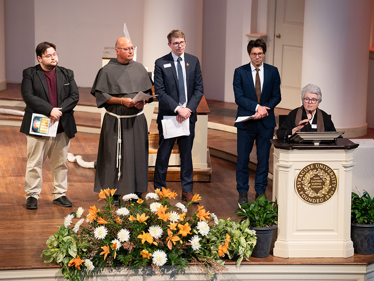 "Five people stand on stage at a Syracuse University formal ceremony while a woman with gray hair speaks at the university podium, flanked by orange and white floral arrangements."