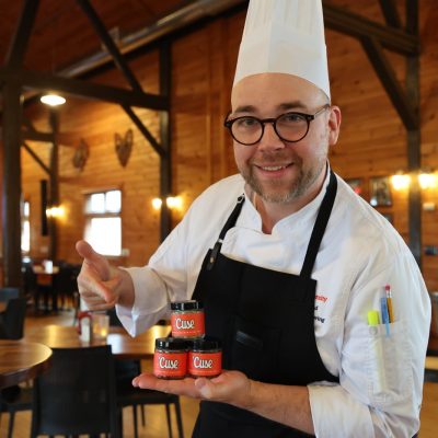 A chef in a white coat, black apron, and tall toque holds three jars of 'Cuse flake salt while giving a thumbs up in a wood-paneled dining hall