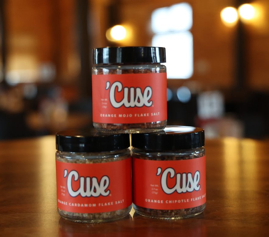 Three small glass jars of 'Cuse branded flake salt — Orange Mojo, Orange Cardamom, and Orange Chipotle — stacked in a pyramid on a wooden table in a warmly lit restaurant setting.