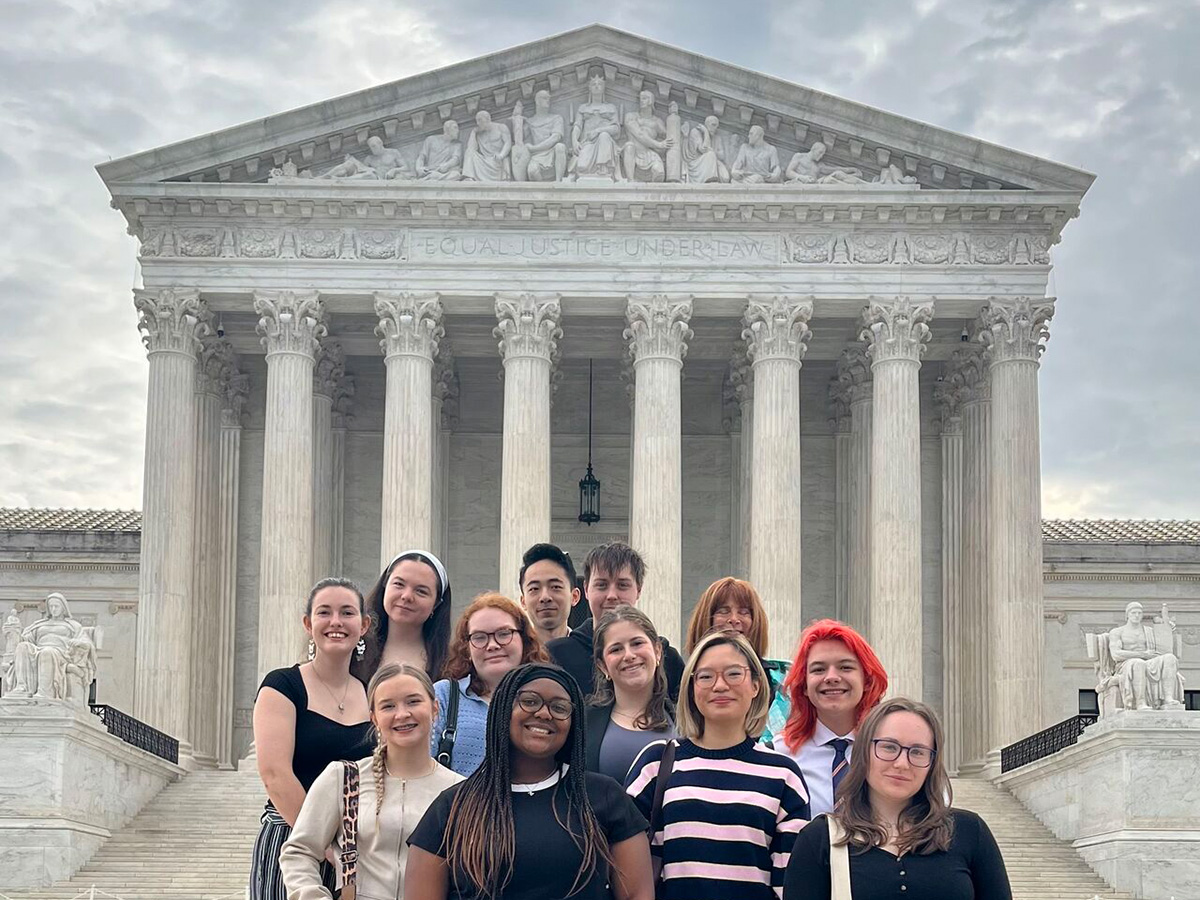A group of approximately 13 students and a faculty member pose on the steps of the U.S. Supreme Court building in Washington, D.C., with the inscription 'Equal Justice Under Law' visible above them