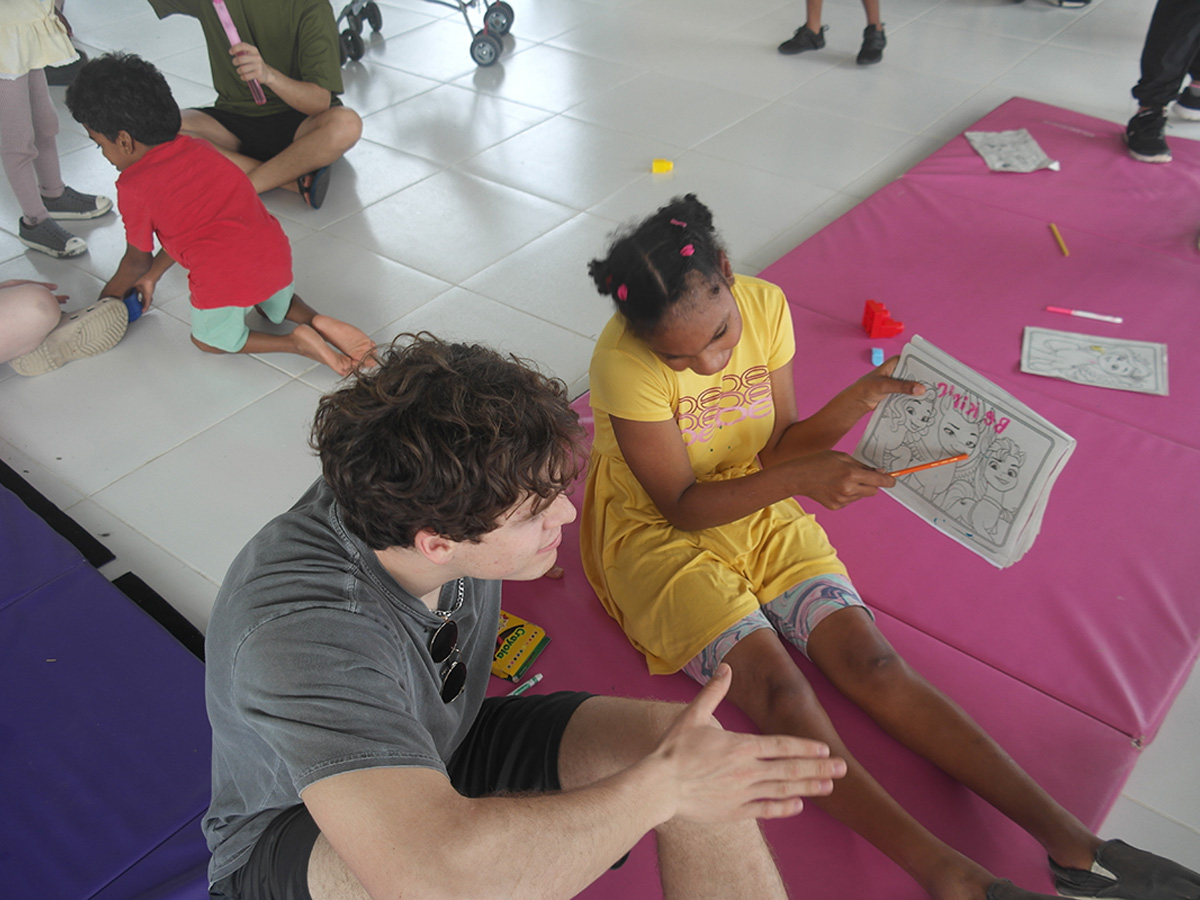 A young adult volunteer sits on a pink mat helping a girl in a yellow shirt with a Barbie coloring page, surrounded by other children coloring on the floor
