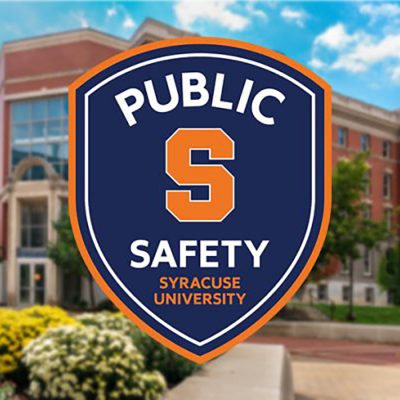 Shield-shaped badge logo for Syracuse University Public Safety, featuring the university's block "S" in orange on a navy blue background with an orange border, displayed over a blurred campus building backdrop.