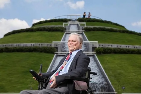 A person seated in a powered wheelchair in front of a large tiered hill with a cascading water feature, under a blue sky with scattered clouds.