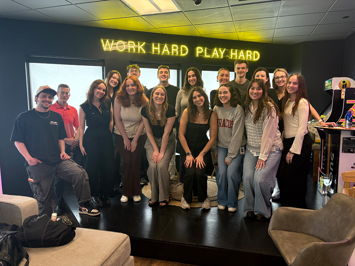 A group of approximately 17 pose together in a modern office lounge beneath a yellow neon sign reading 'Work Hard Play Hard,' with arcade games visible in the background.
