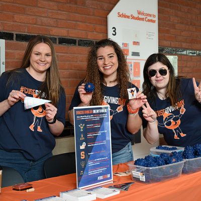 Three smiling Syracuse University staff members or volunteers at a Syracuse Giving Day table inside Schine Student Center, holding promotional items including a mini pennant, a branded ball, and a sticker.