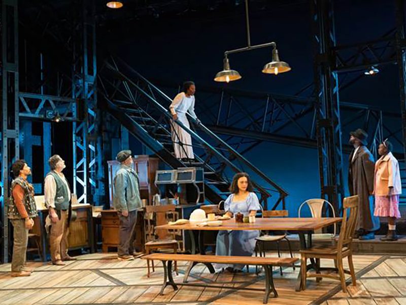 A theatrical stage production featuring seven actors on an industrial set with metal scaffolding, a central staircase, wooden furniture, and warm pendant lighting against a deep blue backdrop."
