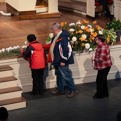 People place white carnations on a stage ledge during a memorial ceremony at Syracuse University. Orange and white floral arrangements decorate the stage, and the Syracuse University seal is visible on a pillar in the upper right. Four attendees are seen in line, each holding or laying a white flower in tribute.