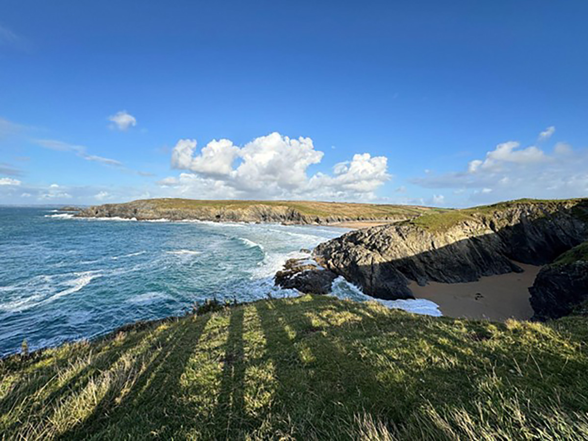 A rugged coastal clifftop view overlooking a sandy cove, with rolling waves, layered rock formations, and green headlands stretching into the distance under a bright blue sky with scattered clouds.