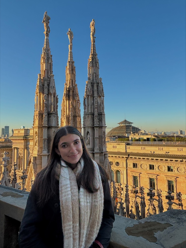 Person standing on a terrace with ornate Gothic spires and a historic stone building in the background at sunset.