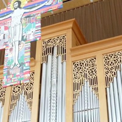 A pipe organ with light wood cabinetry and ornate decorative lattice panels flanking rows of silver metal pipes, installed in a church. To the left hangs a large colorful crucifix banner depicting Jesus on the cross, decorated with the flags of many nations."