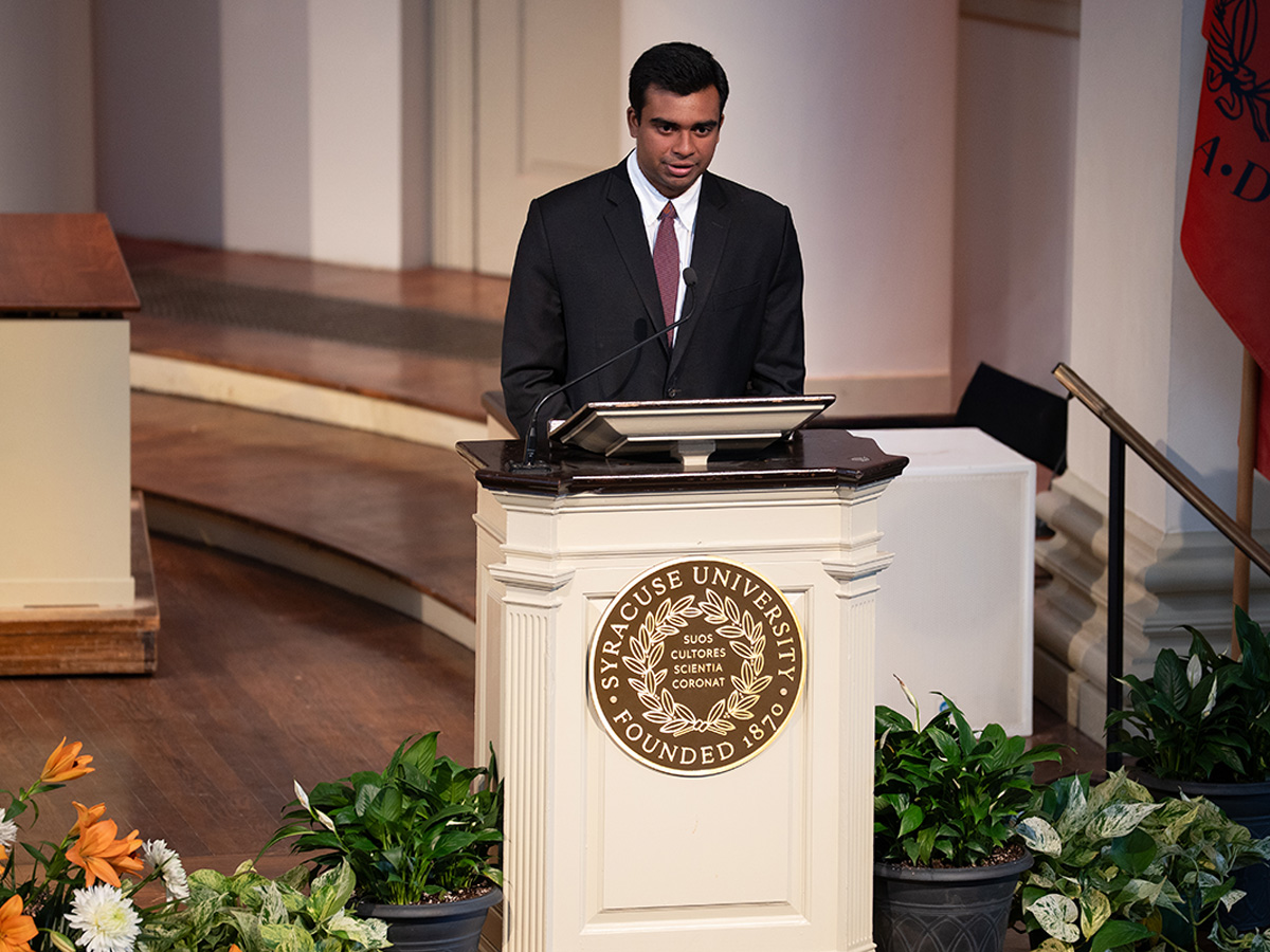 "A man in a dark suit and burgundy tie speaks at a Syracuse University podium during a formal ceremony, surrounded by potted plants and flowers."