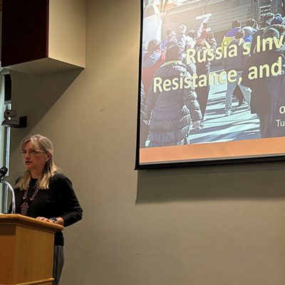 A speaker stands at a wooden podium with a microphone, presenting a lecture titled "Russia's Invasion, Resistance, and Pros[pects]" at an academic venue. A projection screen behind her displays the presentation slide, which credits the speaker as "Oxana Sheve[tsova/li]" from Tufts University. The speaker is a woman with shoulder-length blonde hair and glasses, wearing a black top.