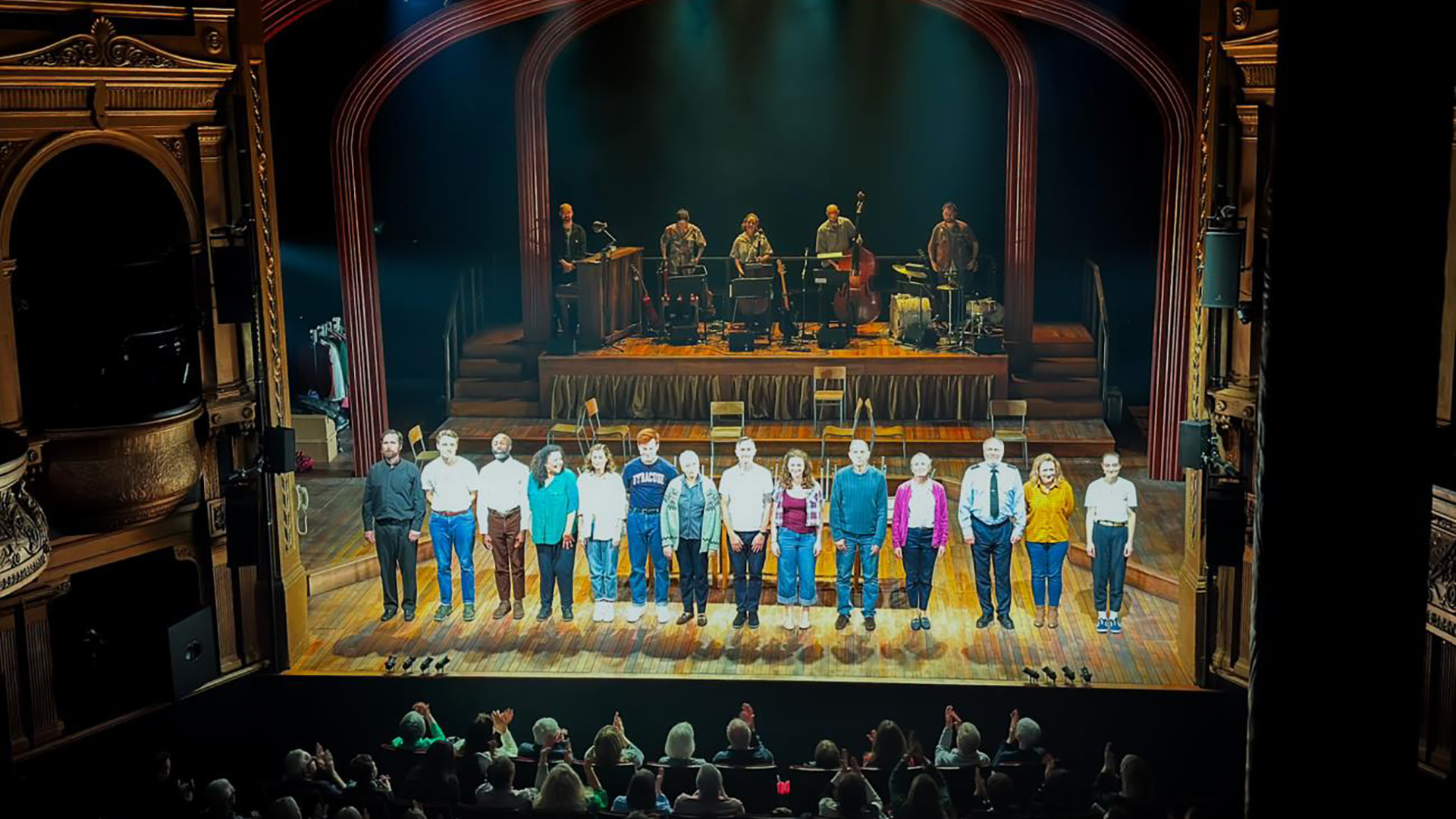 A theater cast takes a curtain call on an ornate stage, lined up in a row while an audience applauds. A live band is visible on a raised platform behind them.