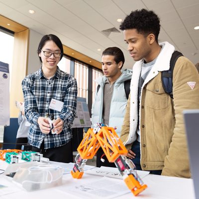 Tom Xiao demonstrates a robotic device to two fellow students at a research symposium display table, with research posters visible in the background.