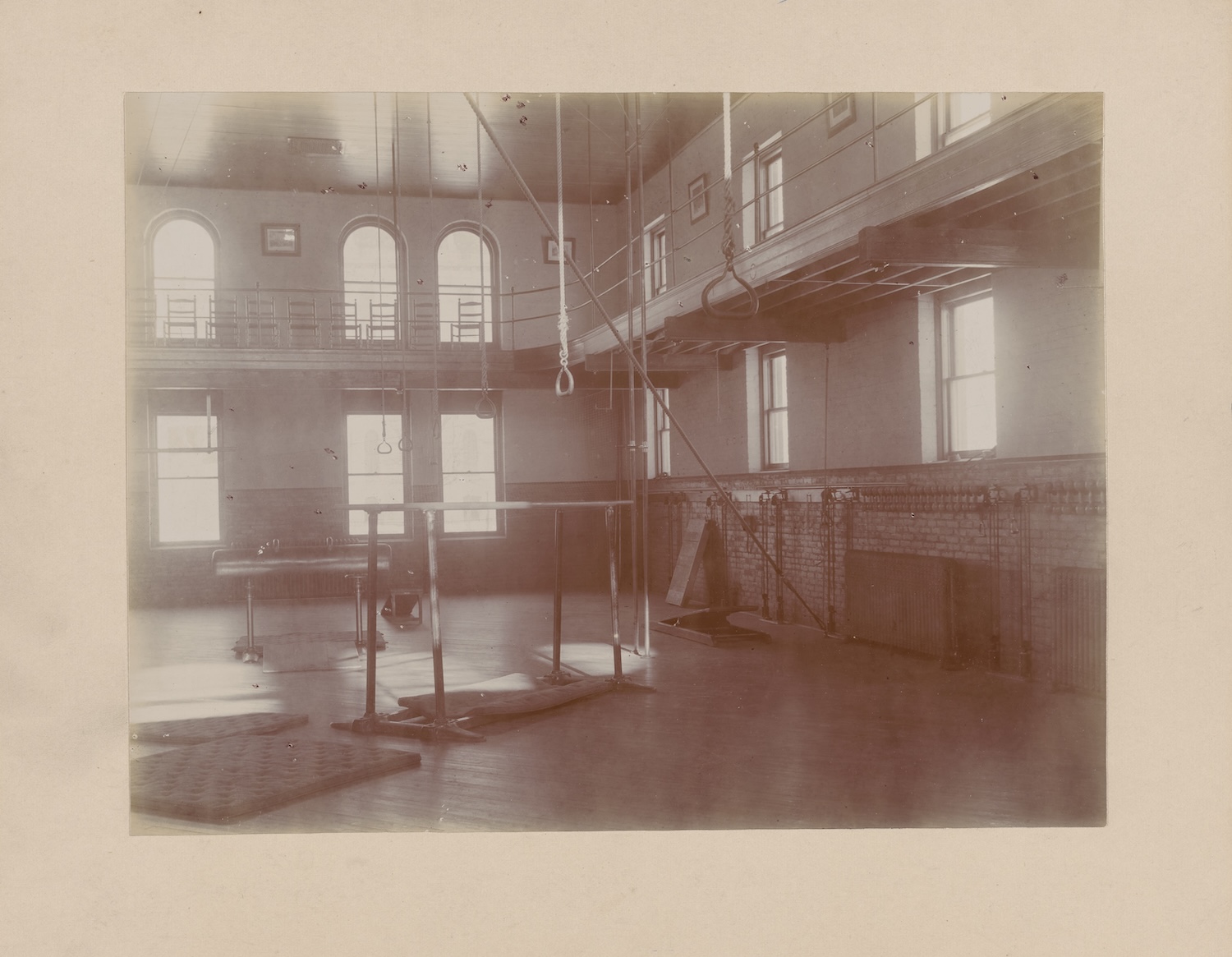 Vintage photograph of an empty gymnasium with wooden floors, ropes, rings, parallel bars, and large arched windows.