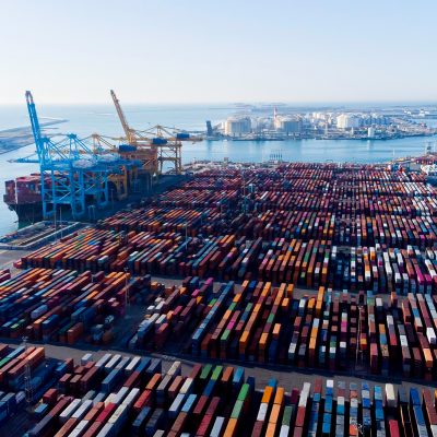 Aerial view of a large commercial shipping port with thousands of colorful intermodal cargo containers stacked in rows across a vast terminal yard, large container cranes along the dock, a cargo ship being loaded or unloaded, and an industrial waterfront cityscape visible across the harbor in the background.