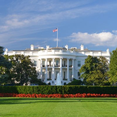 The White House South Lawn, with a fountain, red flower beds, and the American flag flying above the portico on a sunny day.