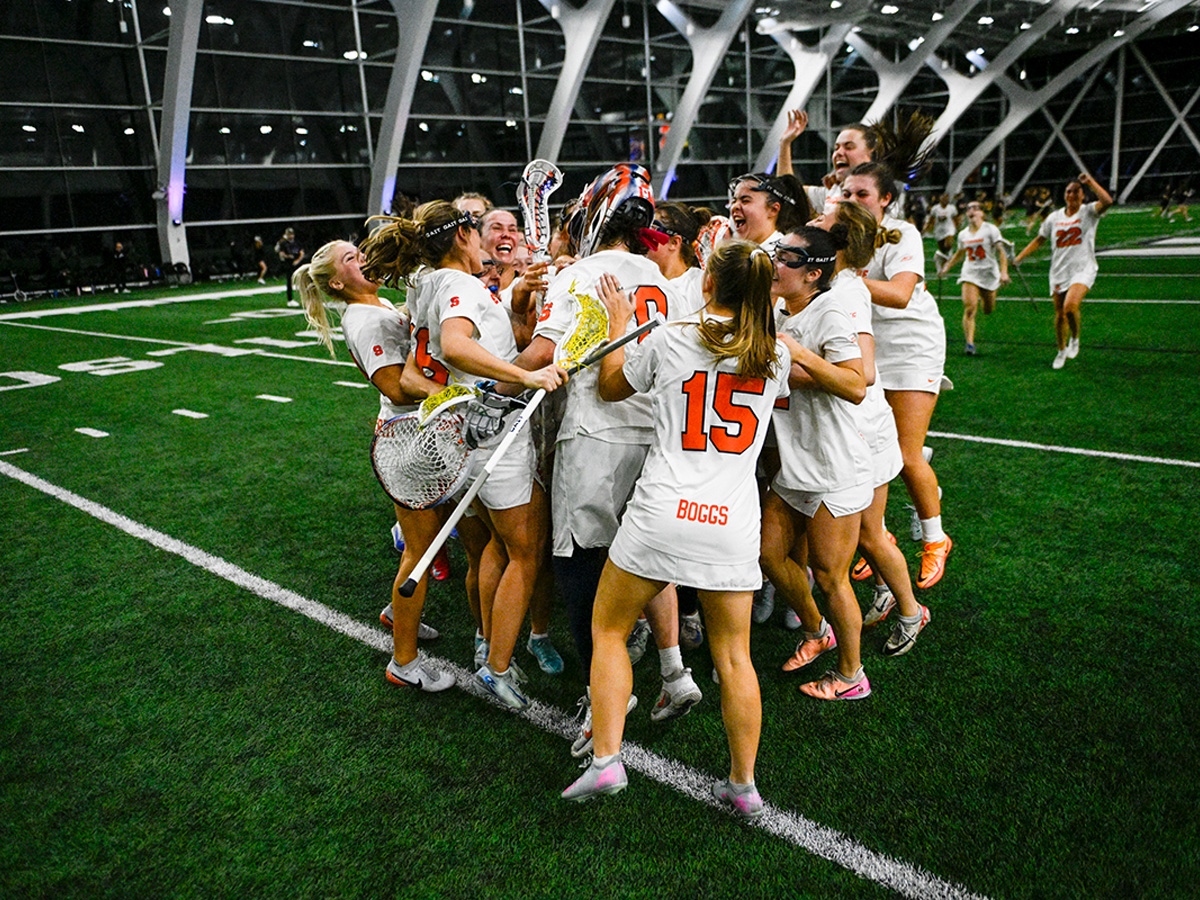 Syracuse University women's lacrosse players celebrate together on an indoor turf field, jumping and embracing in a joyful team huddle after a win