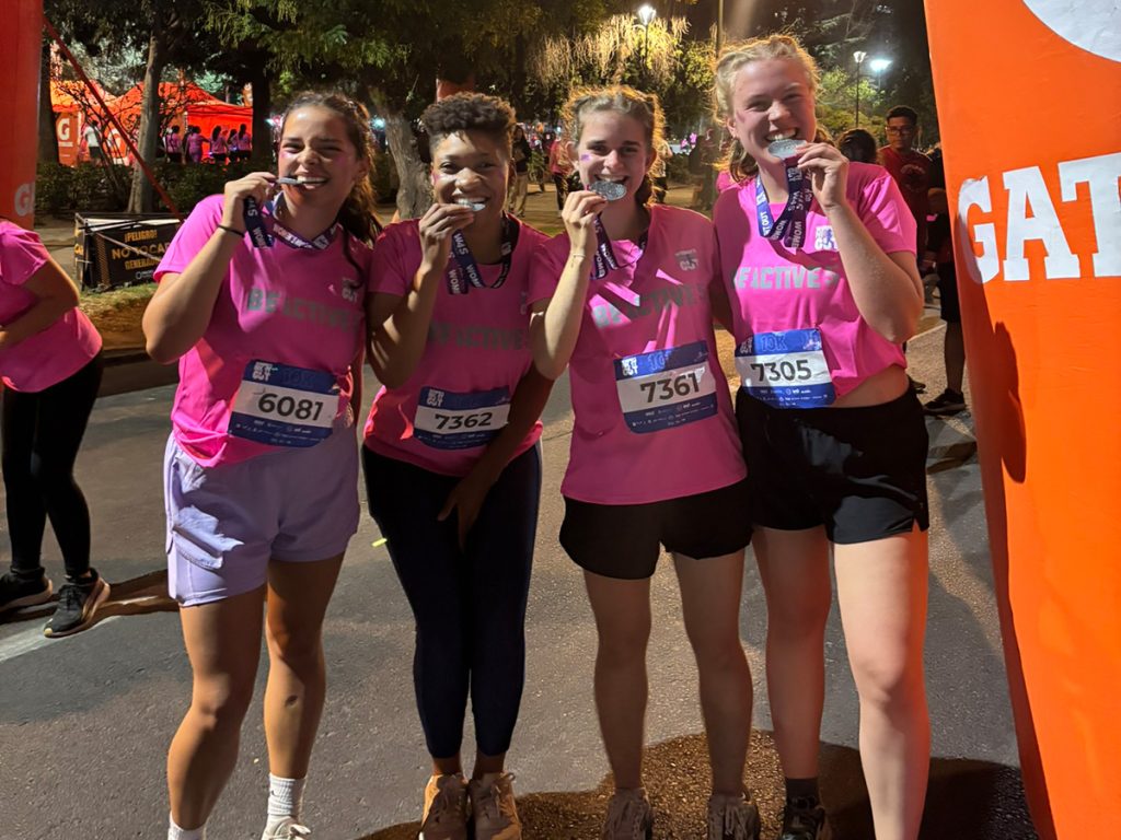 Four women in matching pink "Be Active" t-shirts and race bibs pose together at a nighttime 10K finish line, each playfully biting their finisher medals and smiling at the camera